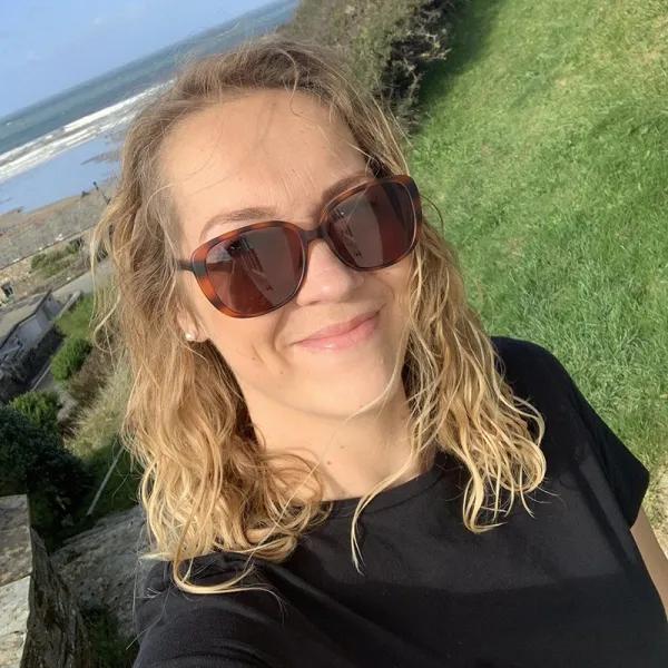 Picture shows a headshot of a lady with mid shoulder dark blonde hair, wearing sunglasses, with green grass over her shoulder, and the sea in the distance behind her. 
